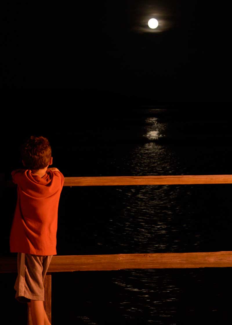 Photograph of a boy watching the full moon rise over water.